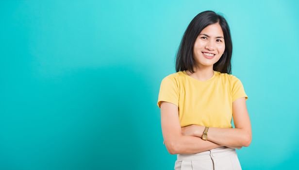 Portrait Asian beautiful young woman standing smile seeing white teeth, She crossed her arms and looking at camera, shoot photo in studio on blue background. copy space to put text on left-hand side.