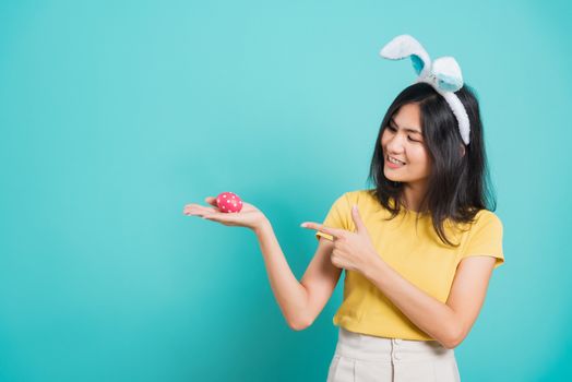 Portrait Asian beautiful happy young woman smile white teeth wear yellow t-shirt standing with bunny ears and holding Easter eggs and point it her looking to camera, on blue background with copy space