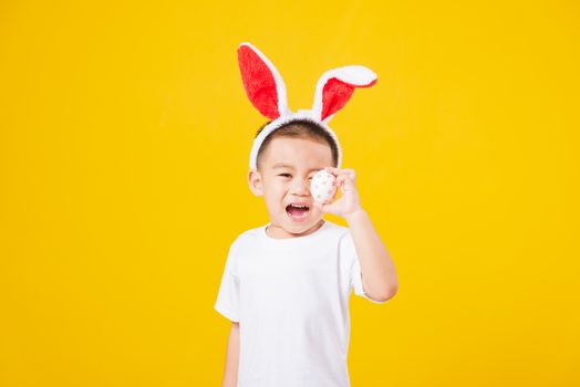 Portrait happy Asian cute little children boy smile standing so happy wearing white T-shirt and bunny ears in Easter festival day holding easter eggs, studio shot on yellow background with copy space