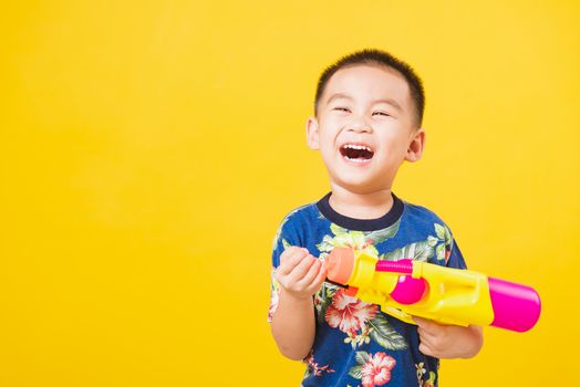 Portrait happy Asian cute little children boy smile standing so happy wearing flower shirt in Songkran festival day holding water gun, studio shot on yellow background with copy space