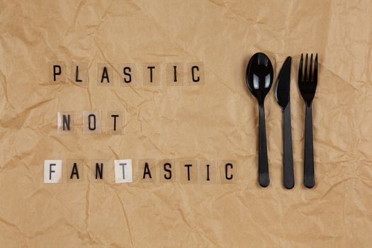 Disposable black appliances fork, spoon, knife, phrase from letters on transparent base Plastic not fantastic on brown crumpled craft paper. Eco, zero waste concept. Flat lay, top view. Horizontal.