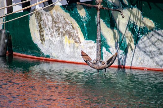The anchor of a grungy boat is seen hanging above rippling water before the boat's hull.