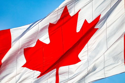 A close-up view of a large Canadian Flag shows the famous red maple leaf symbol on a white background, rippling as it blows in the wind.