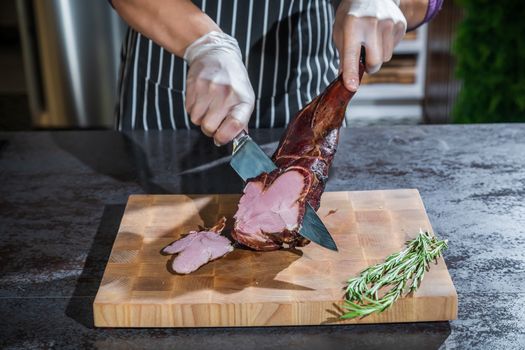 A cook cuts a smoked lamb leg into slices on a wooden cutting board with a knife.