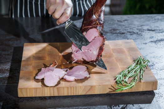 A cook cuts a smoked lamb leg into slices on a wooden cutting board with a knife.