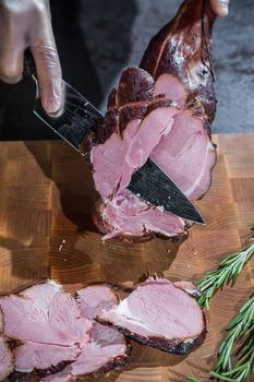 A cook cuts a smoked lamb leg into slices on a wooden cutting board with a knife.