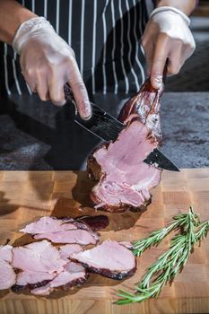 A cook cuts a smoked lamb leg into slices on a wooden cutting board with a knife.