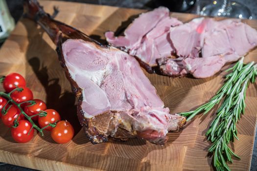 Sliced smoked lamb leg with tomatoes and rosemary on a wooden cutting board.