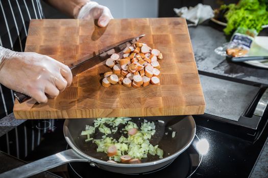 The cook transfers the chopped onion and sausages from the cutting board into a heated frying pan