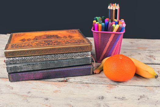 Concept of school lunch break with banana, tangerine and school supplies on wooden desk, selective focus. Books, markers and color pencils emphasizing the concept. Time for study and relax. Copy space. Black background