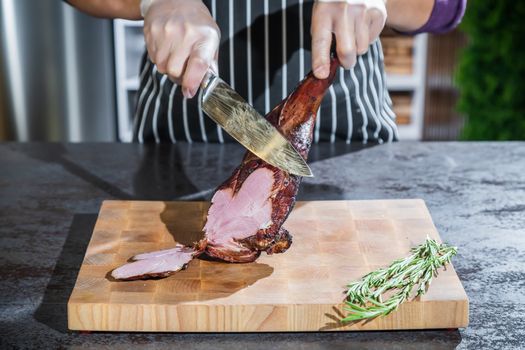 A cook cuts a smoked lamb leg into slices on a wooden cutting board with a knife.