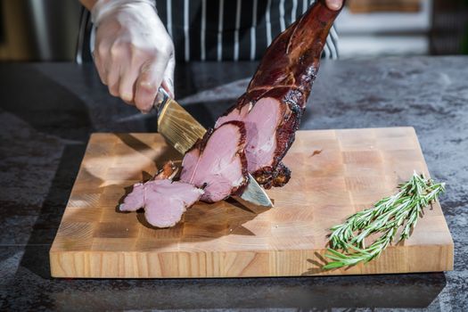 A cook cuts a smoked lamb leg into slices on a wooden cutting board with a knife.