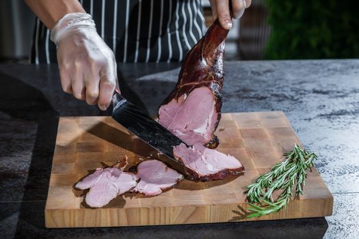 A cook cuts a smoked lamb leg into slices on a wooden cutting board with a knife.