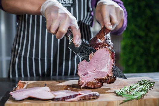 A cook cuts a smoked lamb leg into slices on a wooden cutting board with a knife.