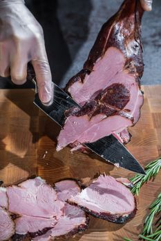 A cook cuts a smoked lamb leg into slices on a wooden cutting board with a knife.