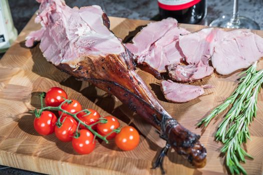 Sliced smoked lamb leg with tomatoes and rosemary on a wooden cutting board.