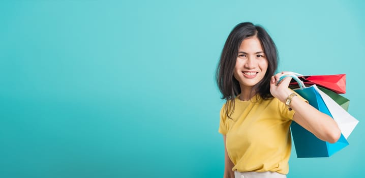 Portrait happy Asian beautiful young woman smile white teeth standing wear yellow t-shirt, She holding shopping bags on hand her looking camera, shoot photo in studio on blue background with copyspace