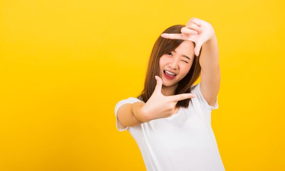 Asian happy portrait beautiful cute young woman standing wear t-shirt hands and finger making frame gesture camera for photography looking to camera, studio shot on yellow background with copy space