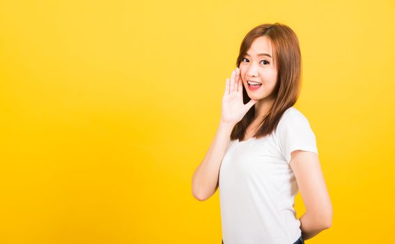 Asian happy portrait beautiful cute young woman teen standing wear t-shirt hand on mouth talking whispering secret rumor looking to camera isolated, studio shot on yellow background with copy space