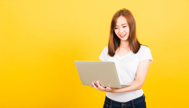 Asian happy portrait beautiful cute young woman teen smiling standing wear t-shirt using laptop computer on hand looking to computer isolated, studio shot on yellow background with copy space