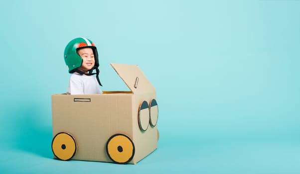 Happy Asian children boy with Helmet smile in driving play car creative by a cardboard box imagination, summer holiday travel concept, studio shot on blue background with copy space for text