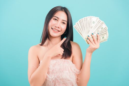 Asian happy portrait beautiful young woman standing smile her celebrating holding dollar money fan banknotes on hand and looking to camera on blue background with copy space for text