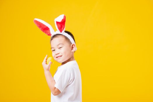 Portrait happy Asian cute little children boy smile standing so happy wearing white T-shirt and bunny ears in Easter festival day holding easter eggs, studio shot on yellow background with copy space