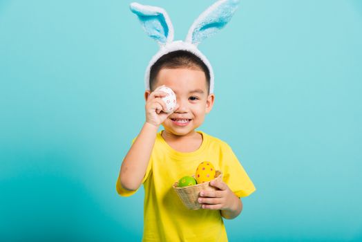 Asian cute little child boy smile beaming wearing bunny ears and a yellow T-shirt, standing to holds colored easter eggs instead of eyes on blue background with copy space for text