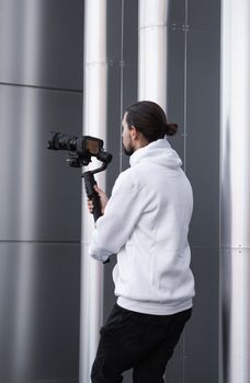 Young Professional videographer holding professional camera on 3-axis gimbal stabilizer. Pro equipment helps to make high quality video without shaking. Cameraman wearing white hoodie making a videos