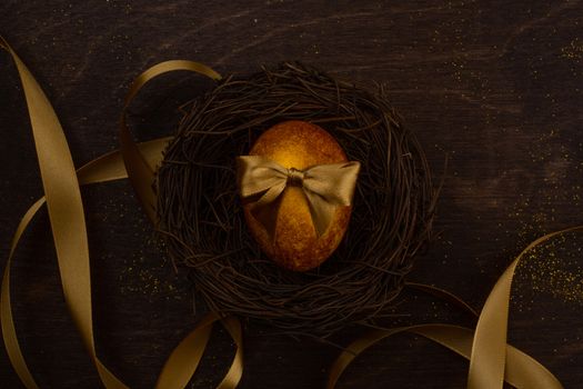 Golden luxury easter egg in nest and ribbon composition on dark wooden background with copy space for text card