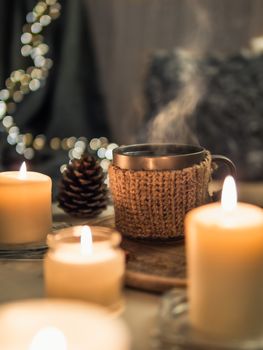 Cozy home, hygge, cosiness concept - hot drink in cup with steam and burning white fragrance candles near sofa with pillows and plaid. Winter decor and hot tea cup. Vertical