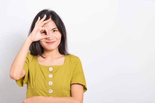 Portrait Asian beautiful young woman standing, She made finger OK symbol sign to agree near eye and looking at camera, shoot photo in studio on blue background, There was copy space