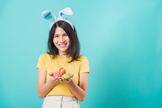 Portrait Asian beautiful happy young woman smile white teeth wear yellow t-shirt standing with bunny ears and holding Easter eggs looking to camera, on a blue background with copy space