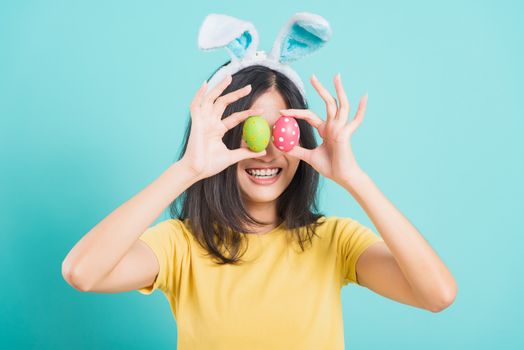 Portrait Asian beautiful happy young woman smile white teeth wear yellow t-shirt standing with bunny ears and holding Easter eggs near the eye, on blue background with copy space