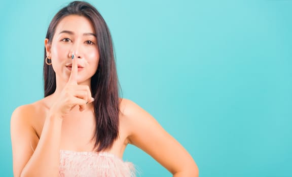 Smiling face Asian beautiful woman her asking quiet with finger on lips pointing with hand on blue background, with copy space for text