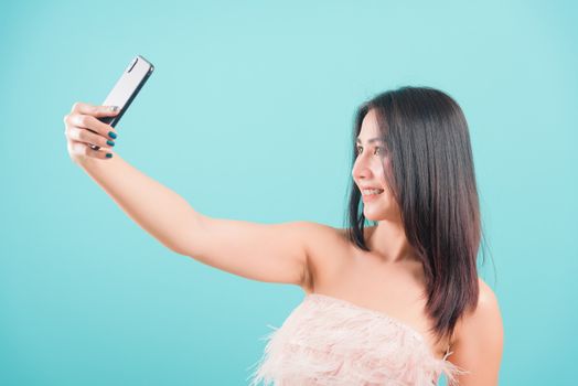 Asian happy portrait beautiful young woman standing smiling her take a selfie video-call on front camera smartphone on blue background with copy space for text