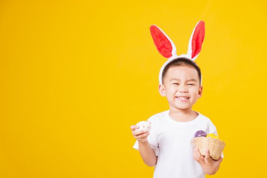 Portrait happy Asian cute little children boy smile standing so happy wearing white T-shirt and bunny ears in Easter festival day holding easter eggs, studio shot on yellow background with copy space