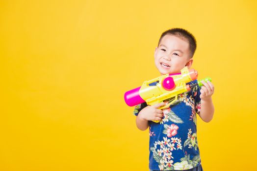 Portrait happy Asian cute little children boy smile standing so happy wearing flower shirt in Songkran festival day holding water gun, studio shot on yellow background with copy space