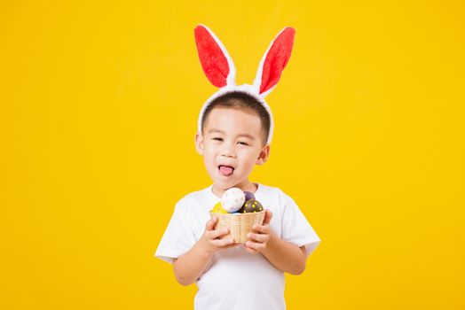 Portrait happy Asian cute little children boy smile standing so happy wearing white T-shirt and bunny ears in Easter festival day holding easter eggs, studio shot on yellow background with copy space