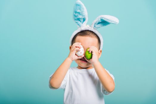 Asian cute little child boy smile beaming wearing bunny ears and a white T-shirt, standing to holds colored easter eggs instead of eyes on blue background with copy space for text