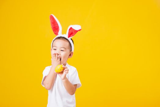 Portrait happy Asian cute little children boy smile standing so happy wearing white T-shirt and bunny ears in Easter festival day holding easter eggs, studio shot on yellow background with copy space