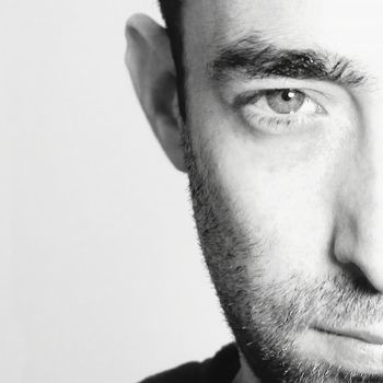 Dramatic closeup portrait of a young bearded man half expressionless face on white background looking at the camera. He is with serious facial expression. Black and white toned image.