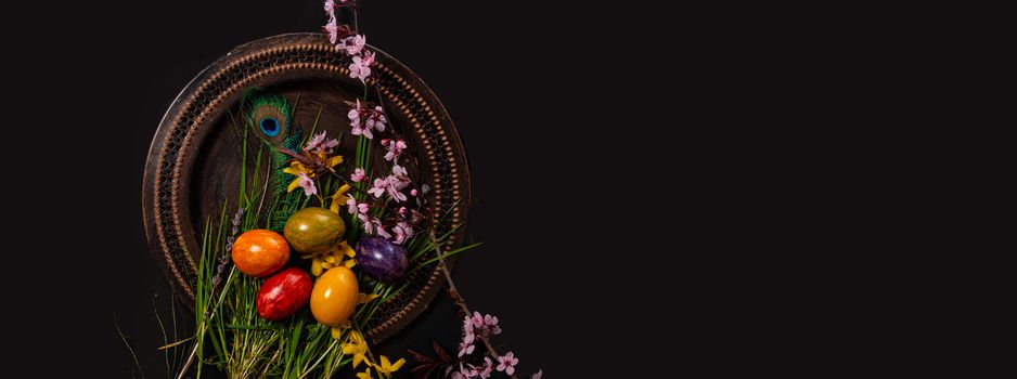 Colored easter Eggs in a plate with black background and copy space. With grass and flowers