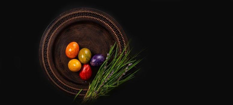 Colored easter Eggs in a plate with black background and copy space. With grass and flowers