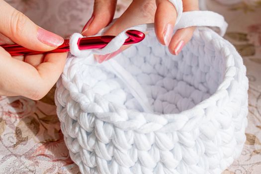 Young woman while crocheting, close up. Stay at home leisure activity idea. White basket made of white T-shirt yarn, with red crochet needle. Pale pink nails.