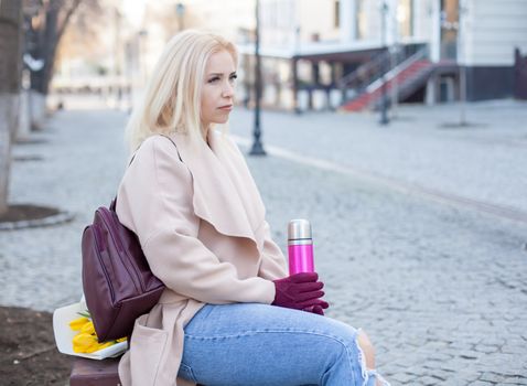 Outdoor of gorgeous woman in elegant coat and gloves sitting in street with thermos of hot tea