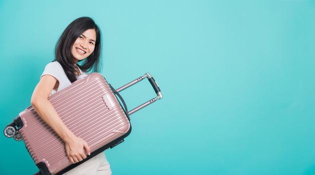 Portrait happy Asian beautiful young woman standing wear white t-shirt, holidays travel concept, her hugging suitcase bag and looking to camera, shoot photo in a studio on a blue background