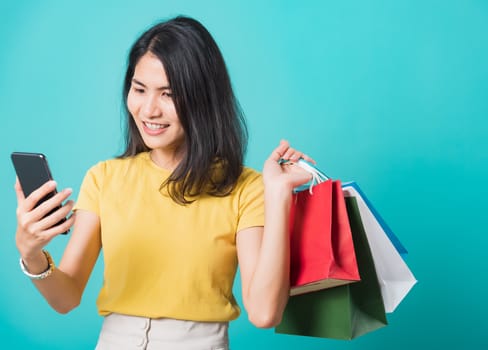 Portrait happy Asian beautiful young woman smile white teeth standing wear yellow t-shirt, She holding shopping bags and using a mobile phone, studio shot on blue background with copy space for text