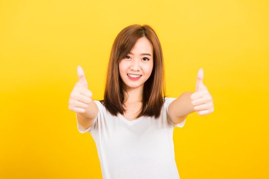 Asian happy portrait beautiful cute young woman teen standing wear t-shirt showing gesturing finger thumb up looking to camera isolated, studio shot on yellow background with copy space