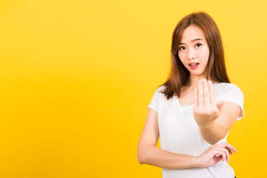 Asian happy portrait beautiful cute young woman teen smile standing wear white t-shirt making gesture hand inviting to come looking to camera isolated, studio shot on yellow background with copy space